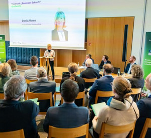 Blick von hinten über die Reihen der Teilnehmenden. Im Hintergrund vor einen großen Leinwand steht Ministerin Ahnen am Rednerpult.