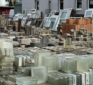 Das Bild zeigt ein Materiallager mit verschiedenen Baustoffen. Im Vordergrund befindet sich ein ordentlich gestapelter Block aus Glasbausteinen. Die einzelnen Glassteine sind quadratisch, durchsichtig bis milchig und haben eine strukturierte Oberfläche. Sie sind gleichmäßig übereinander gestapelt und bilden einen kompakten Stapel.  Im Hintergrund sind mehrere ausgebaute Fenster zu sehen. Die Fensterrahmen stehen oder lehnen hintereinander. Es handelt sich um unterschiedliche Größen und Ausführungen. Einige Rahmen wirken weiß oder hell, andere dunkler. Die Glasscheiben sind teilweise noch eingesetzt.  Die Umgebung erinnert insgesamt an ein Lager oder einen Außenbereich, in dem gebrauchte Baumaterialien gesammelt und aufbewahrt werden.